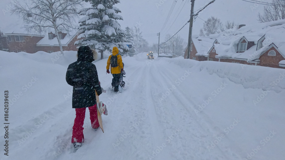 Naklejka premium Two snowboarders walk along the snow-covered road leading through ski resort town in Japan. Snowboarders weather the snowstorm returning from the slopes of Niseko, walking through the quiet town.