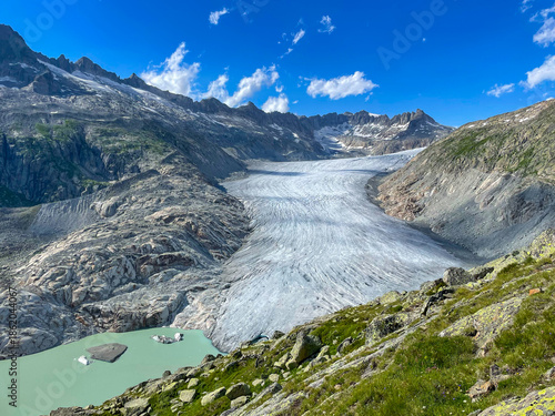 Fotografía Massive Rhone Glacier winds through a rugged mountain landscape under bright blue skies, culminating in a stunning turquoise alpine lake