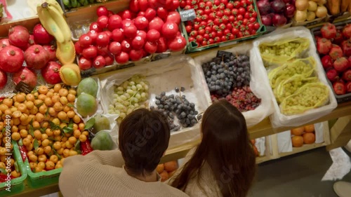 Couple selecting fresh produce, cozy indoor market stall with colorful fruits and vegetables, man in beige sweater and woman in cream sweater browse pomegranates, grapes and tomatoes Top view