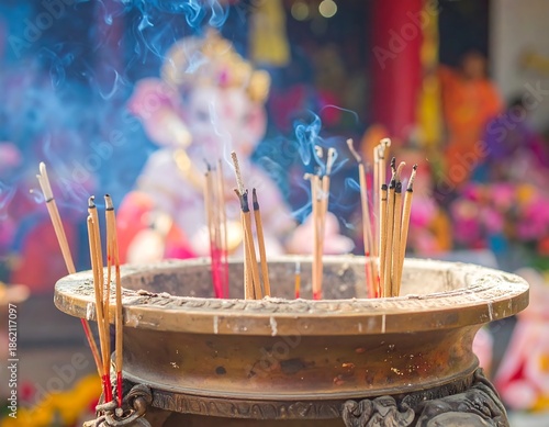 Incense sticks burning in a large pot, creating smoke during a religious ceremony or offering, with a blurred background of a temple interior.