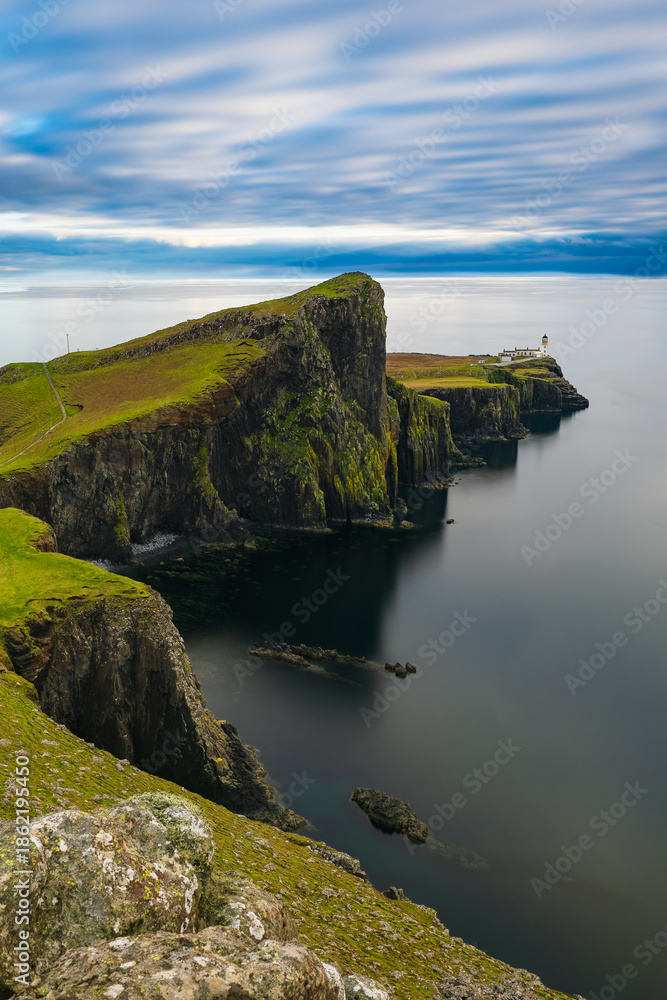 Obraz premium Vertical view of Neist Point cliffs and lighthouse on the Isle of Skye, Scotland, calm ocean and dramatic coastline under cloudy sky, iconic travel landscape with copy space.