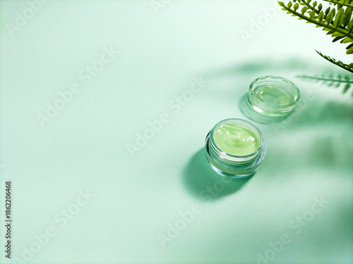 A transparent cosmetic jar with a green cream sits open on a mint-green surface.