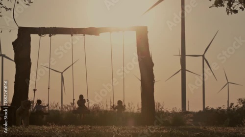 silhouette video of several children playing on swings, with wind turbines in the background.
