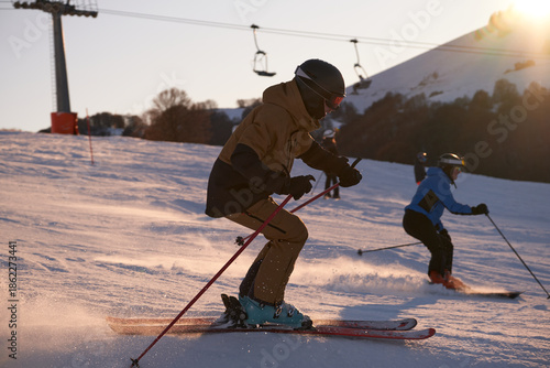 Side view of a skier in a brown suit descending a ski slope at high speed. Alpine skis cut through snow spray at sunset in the Italian Alps with sun rays and blue sky.