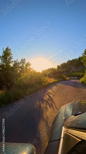 LENS FLARE, VERTICAL: Low morning sun casts golden glow and long shadows on a winding coastal road. Unique perspective of black car traveling through scenic Mediterranean landscape on island of Hvar.