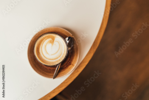 Top Down View of Latte with Heart Foam in Glass Cup on White and Wooden Cafe Table, space for text.