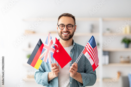 Photos Handsome young guy holding bunch of diverse flags and smiling at camera indoors