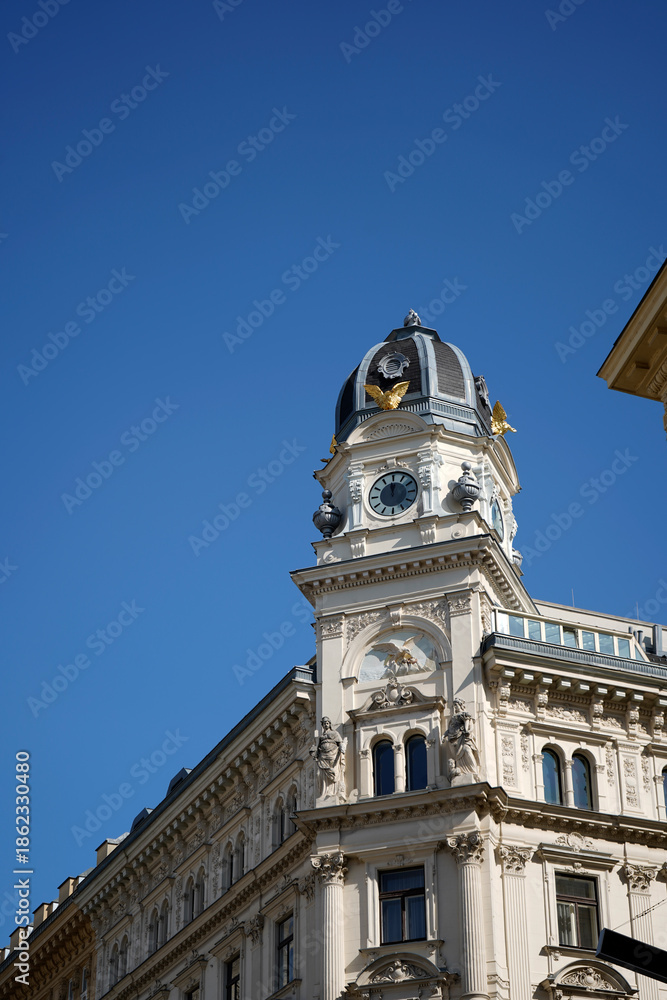 Obraz premium Historic clock tower with ornate facade and domed roof rising against clear blue sky, detailed classical architecture emphasizing time symbolism, urban heritage, and timeless city landmark presence.