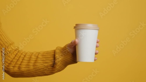 A hand, clad in a mustard sweater, presents a white coffee cup against a yellow background
