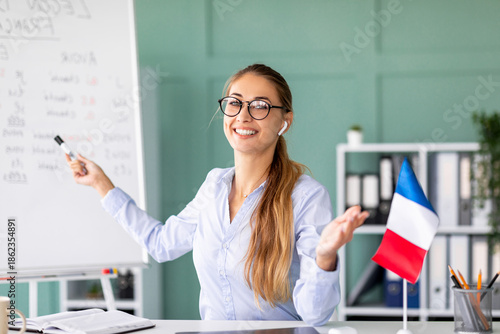 Fotografie A joyful young professor teaches a French class online, pointing at the blackboard that displays essential grammar rules