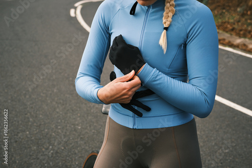 Close-up of a female cyclist getting ready, meticulously putting on her protective gloves on an outdoor asphalt road. She is dressed in blue athletic gear, highlighting focus and readiness for a ride.