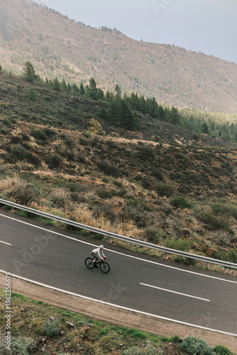 A lone cyclist rides a black road bike along a winding mountain road, surrounded by rugged hillsides and sparse pine trees. Emphasizes endurance, outdoor activity, and scenic travel.