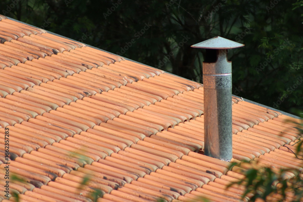 Naklejka premium Close-up of a terracotta tiled roof with a metal chimney pipe and lush green foliage in the background