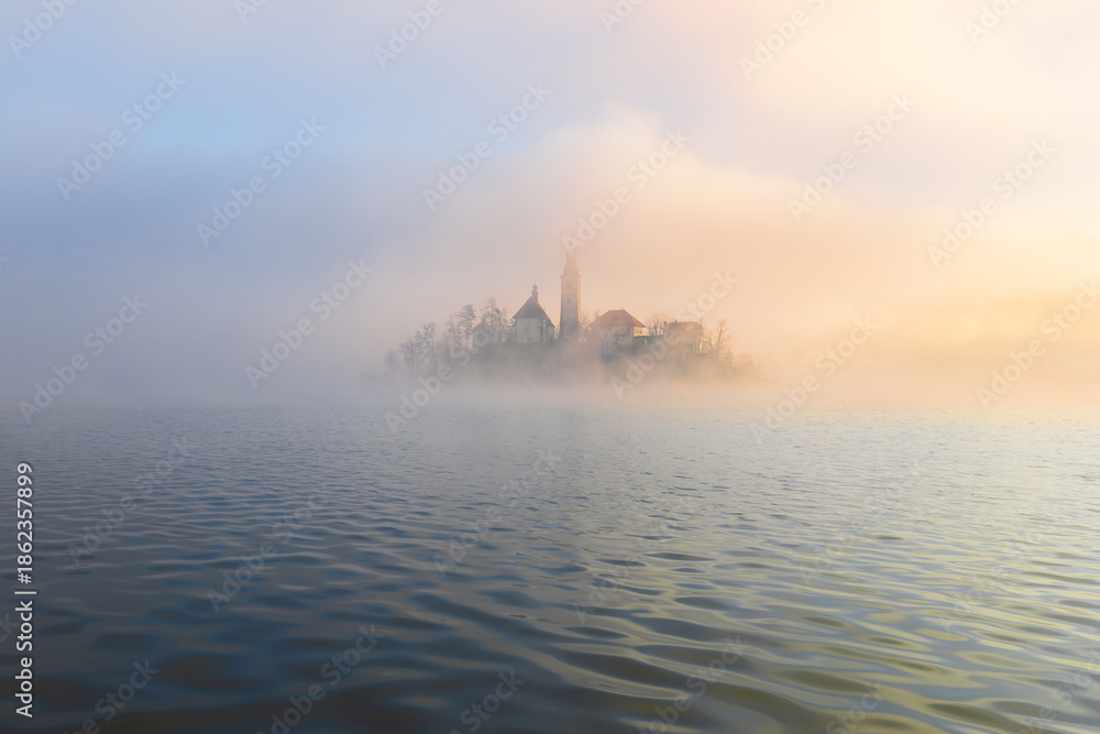 Naklejka premium Mariä Himmelfahrt Kirche auf der Insel bei Bled im dichten goldenen Nebel bei Sonnenaufgang, weite Wasserfläche im Vordergrund und nebelige Landschaft im Morgenlicht in Slowenien