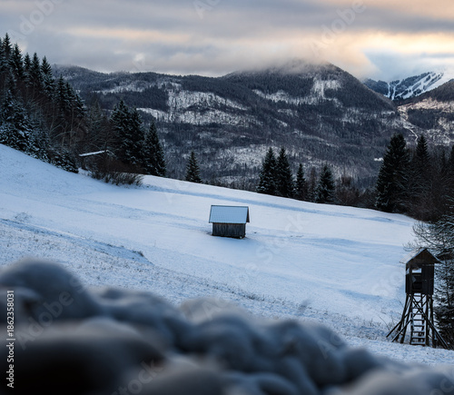 Verschneite Winterlandschaft mit kleiner Hütte auf schneebedeckter Wiese, strukturierter Vordergrund aus Schnee, bewaldete Hügel und Berge im Hintergrund bei Dämmerung und Winterlicht