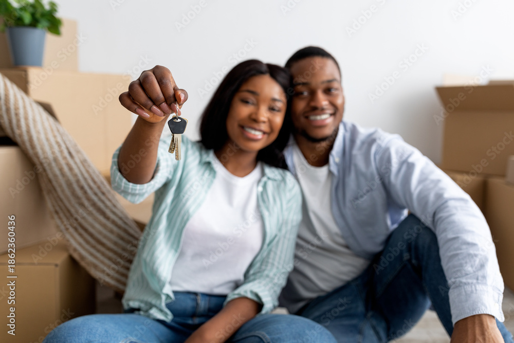 Fototapeta premium Excited black married couple showing home keys while sitting among cardboard boxes, selective focus. Happy spouses celebrating moving to new apartment, enjoying relocation