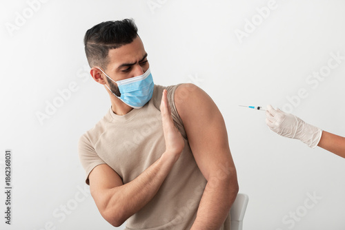Fotografi Young Arab guy in face mask stopping doctor from making covid vaccine injection, being against coronavirus immunization on white studio background