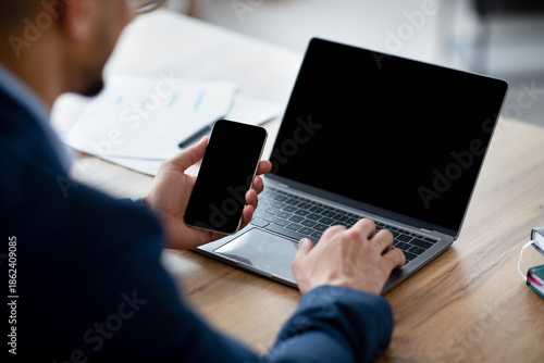 Cropped view of Arab businessman using laptop computer and smartphone with mockup for app or website design, closeup. Unrecognizable male CEO working with modern gadgets, space for text