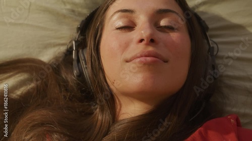 Trendy woman listening to music on old school headphones laying on bed in summer sun - close up