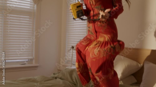 Young Dreamcore woman listing to retro music cassette player jumping up and down on bed