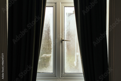 Condensation forming on double-glazed window with closed dark curtains in winter light.