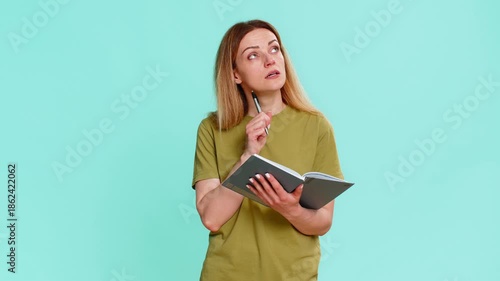 Middle-aged woman writes notes in notebook, pauses with thoughtful expression and looks up, reflecting on life direction or next step. Girl on blue background shows deep concentration and planning