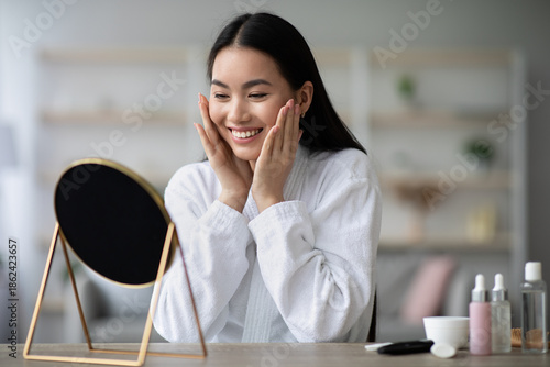 Pretty asian woman in bathrobe making face-lifting massage, sitting at table in bedroom, loking at mirror and smiling, beautiful chinese lady using moisturizing face cream after shower, copy space