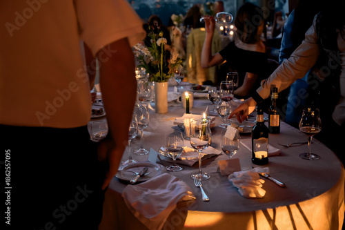 Fotografie Elegant candlelit dinner table at a luxury evening event with wine glasses, floral centerpiece, and guests mingling, capturing a warm, intimate, and sophisticated celebration atmosphere