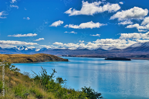 Turquoise lake surrounded by mountains and rolling hills under a blue sky with fluffy clouds. Lake Tekapo with Motuariki island and the snow-capped Southern Alps, South Island, New Zealand
