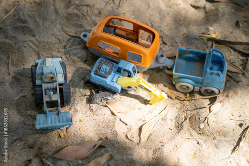 Colorful plastic toys scattered on sandy ground, including blue truck, yellow excavator, and orange vehicle. These toys evoke childhood memories and outdoor play