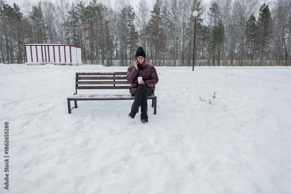 Fototapeta premium solitary woman sitting on snowy bench gazing into distance, wide winter landscape with sparse trees and modern building backdrop, mood of contemplation and quiet pause, footprints in snow and muted