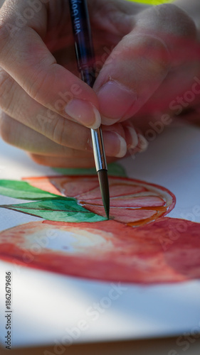 Watercolor painting process showing a hand holding a brush to paint fruit. Beautiful orange and apple illustration being created in an art studio for a creative hobby or tutorial.