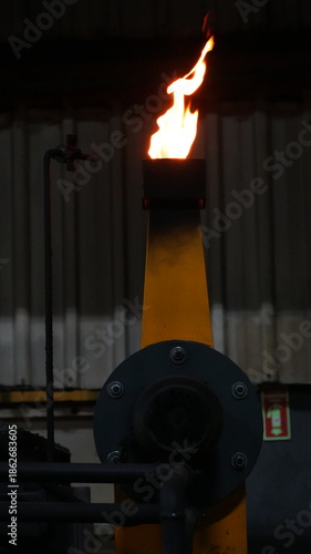 Industrial gas burner with a bright orange flame in a dark factory. Professional equipment for metal forging and heat treatment processes in heavy industry manufacturing.