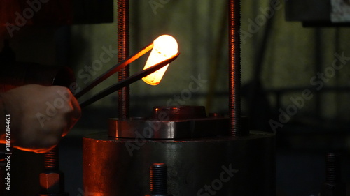 Hand holding tongs with glowing metal piece, intense forge heat, mechanical grip, industrial tool, manufacturing process, red-orange illumination, precision metalwork, workshop atmosphere.