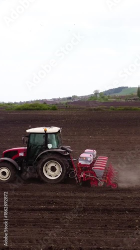 Red tractor plowing a field with a seed drill, showcasing the continuous action of planting seeds in rich soil, with dust rising from the ground