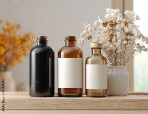 Three amber glass bottles with blank white labels sit on a light-brown wooden surface with dried flowers