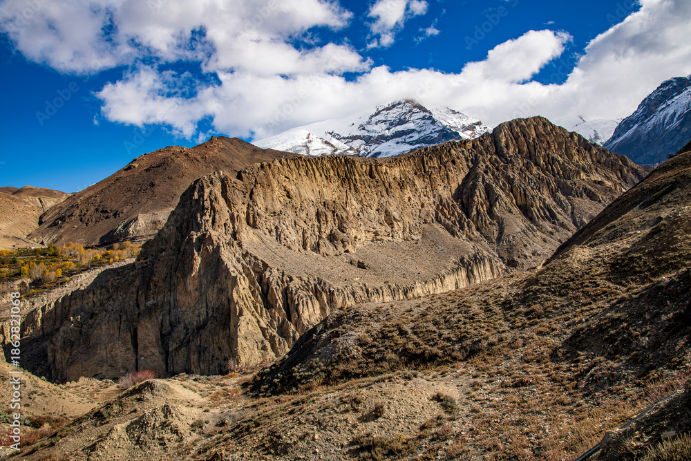 Fototapeta premium Eroded cliffs and ancient cave formations along the trekking route from Muktinath to Kagbeni in the Kali Gandaki Valley, Nepal. The arid, rugged landscape of the Mustang region contrasts with snow-cov