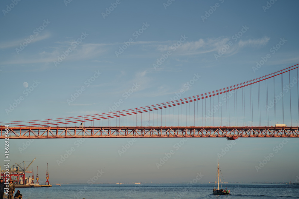 Obraz premium A red suspension bridge spans across the image with a clear blue sky and visible moon.