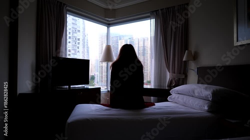 Tired business woman sit dressed on bed at dark room, stand up and go to window, looking out to city. Pensive mood, bedroom illuminated by natural light from one window