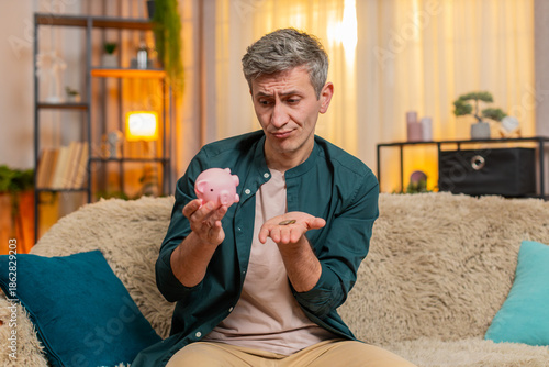 Young man at home opens piggy bank and finds only few coins inside, reacting with disappointment and sadness. Caucasian guy on sofa sighs and shows poor financial condition with quiet frustration.