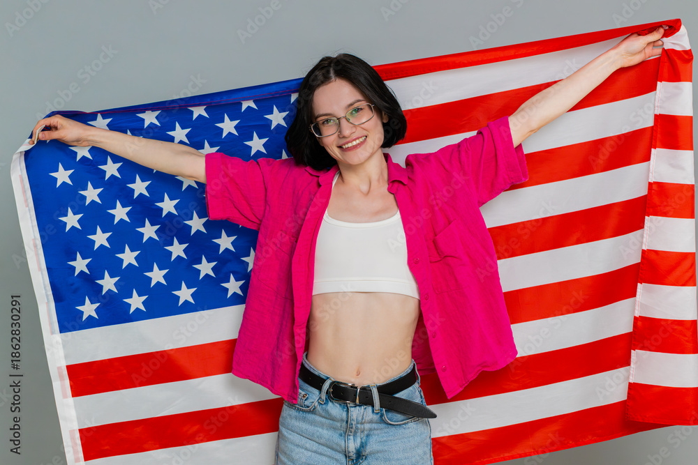 Fototapeta premium Woman happily holds waves American USA flag, proudly showing patriotic emotion or supportive attitude. Caucasian girl demonstrates cheerful national pride and enthusiasm, isolated on gray background