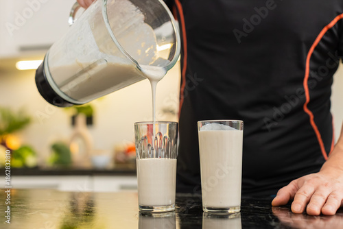 Man pours protein smoothie from blender into glass on kitchen counter for healthy nutrition. Shake with banana milk cottage cheese and whey protein supports muscle recovery weight control fitness gym.