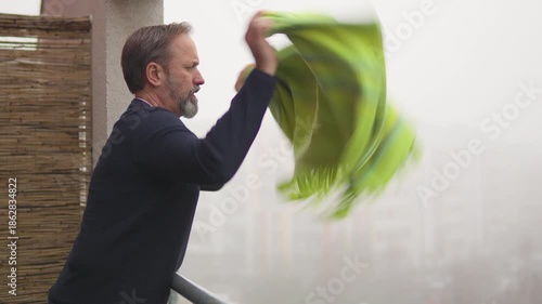 A handsome middle-aged man shakes out a blanket over a railing on a balcony of an apartment