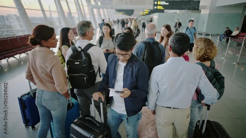 Young man and diverse group of travelers waiting in line at an airport terminal at sunset, checking smartphones and boarding passes for international flight and travel