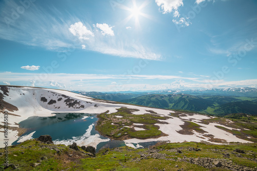 Scenic sunlit landscape with alpine lake in rocky snowy cirque near stone hill top in sunny day during thaw. Ice floats in mountain lake among rocks with view to forest mountain range under bright sun