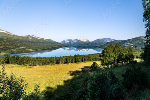 View over a meadow and Lake surrounded by forests with Trollheimen mountains in the Background. Summer blue sky with bright sunlight and clear reflections on the water surface. Oppdal Norway