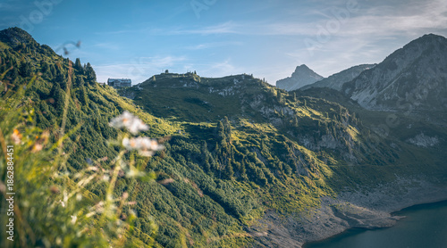 Alpine lake with a traditional mountain hut in a scenic late summer evening landscape, surrounded by peaks, meadows, and forests in soft warm light.