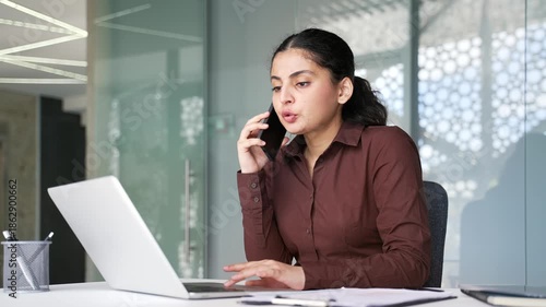 Busy businesswoman multitasking talking on smartphone and working on a laptop while sitting in modern office workplace. Young female employee having business calls, manages tasks, using computer.
