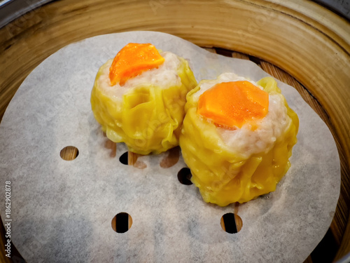 A close-up shot of two yellow-wrapped pork dumplings ,Shumai, topped with bright orange salted egg yolk, served on parchment paper inside a traditional bamboo steamer