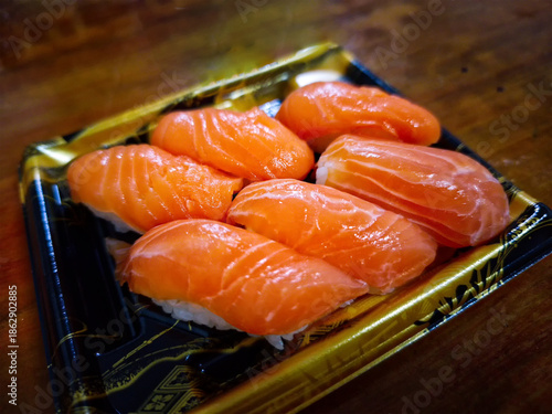 A set of six fresh salmon nigiri sushi pieces with vibrant orange textures, neatly arranged in a decorative black and gold plastic takeout container on a wooden surface